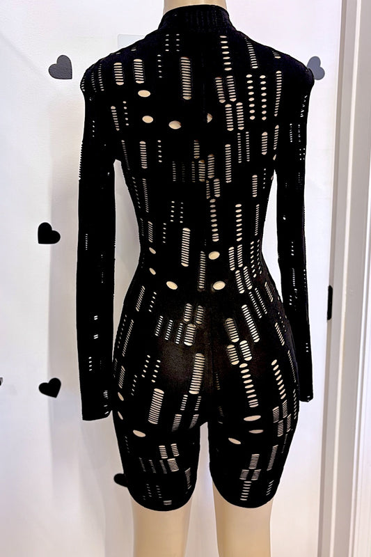 A black long-sleeve knit romper with an all-over cutout geometric pattern, displayed on a mannequin against a white wall decorated with small black heart stickers.