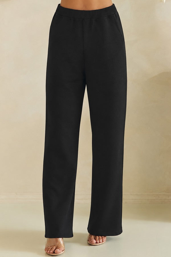 Black wide-leg pants shown on a model from the waist down, featuring a relaxed straight-leg fit and side pockets, paired with neutral-colored flat sandals against a warm beige background.