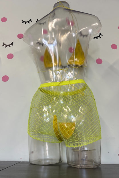 A novelty yellow mesh bikini-style bottom displayed on a clear plastic torso mannequin, with a yellow undergarment visible inside. The mannequin stands on a dark tabletop against a white wall with pink polka dots and black eyelash decals.