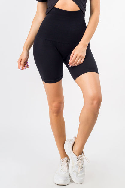 A model wearing black high-waisted biker shorts, a black cutout top, and white sneakers, shown from the waist down against a light gray studio background.