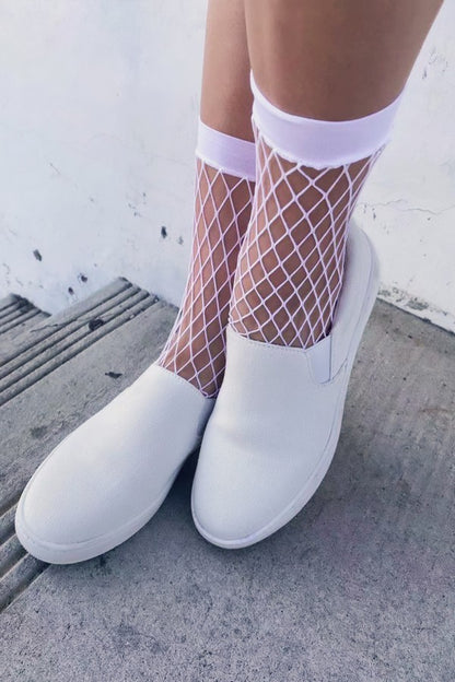 White slip-on shoes worn with white fishnet socks, shown on a person's feet against a gray concrete and white wall background.