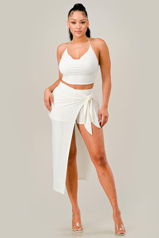 Woman wearing an off white outfit with a halter top and wrap tie skirt on a plain background