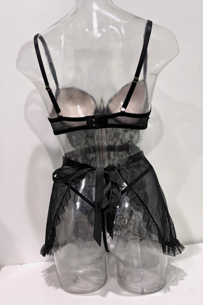 Black lace lingerie set displayed on a clear plastic mannequin torso, featuring a sheer underwire bra with black straps and a matching sheer apron-style skirt with a large front bow.