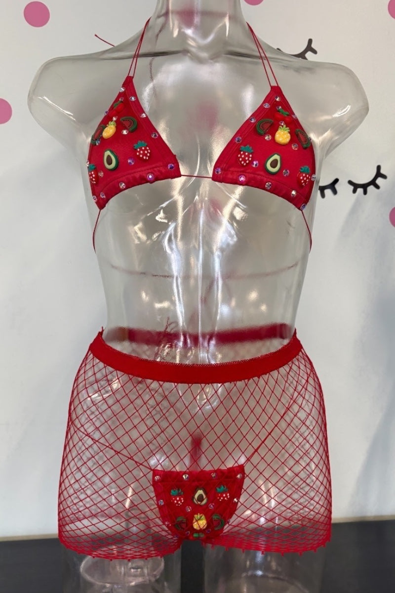 A two-piece red bikini set displayed on a clear plastic mannequin torso. The top is a triangle halter style with a red fabric printed with colorful fruit illustrations, and the bottom are red fishnet mini shorts with a matching red fishnet thong at the front containing the fruit print.