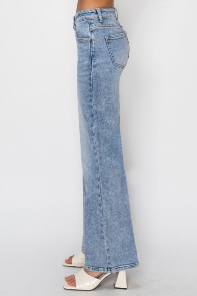 A person shown from the waist down wearing light-wash blue flare jeans and white heeled sandals against a plain light gray studio background.