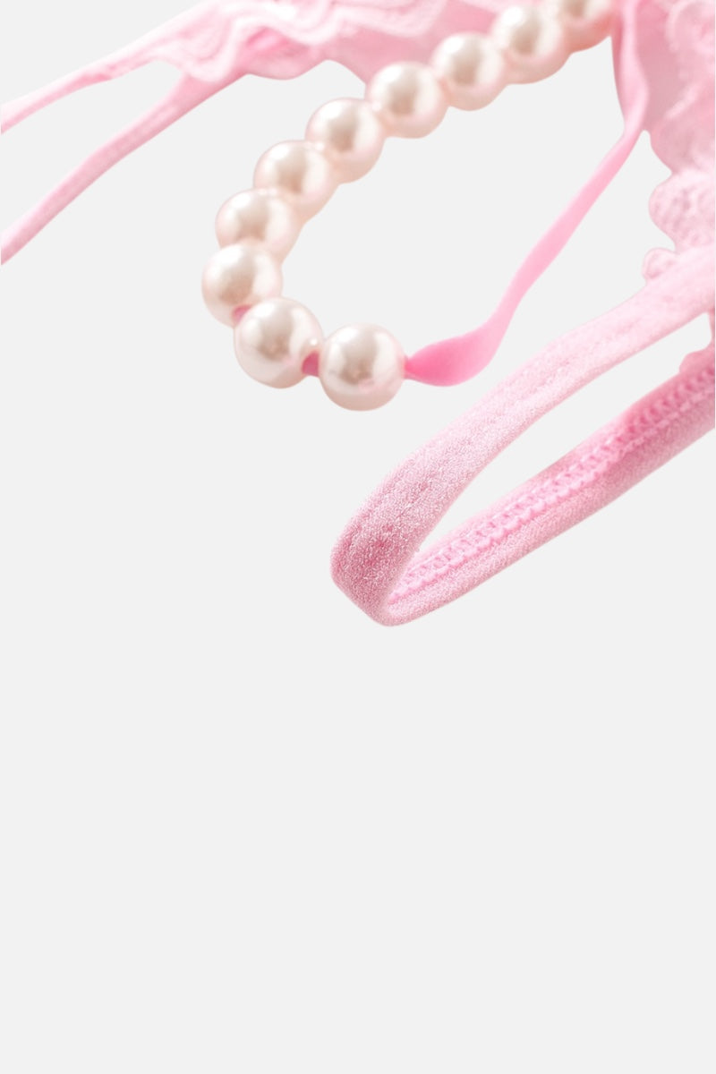 Close-up of a light pink lingerie piece with lace trim and a curved row of white faux pearls, shown against a plain light gray background.