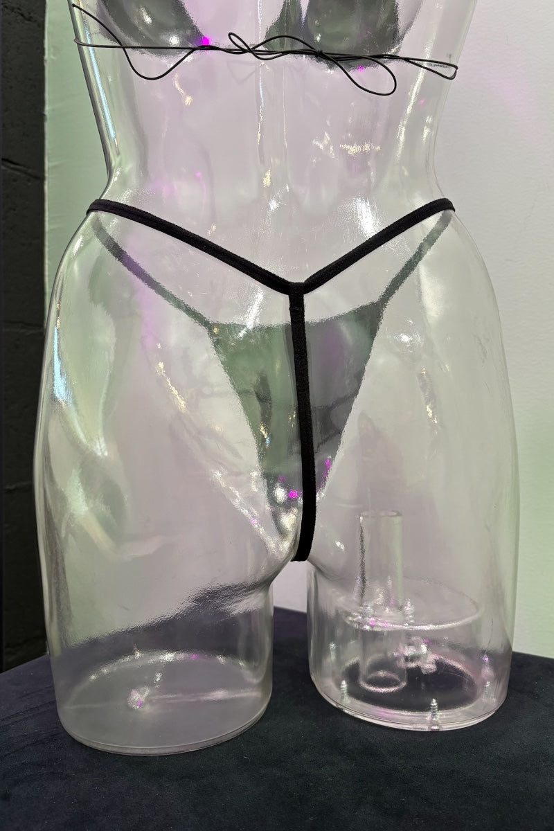 Clear plastic lingerie display form shaped like a torso, wearing black thong-style underwear, with a matching clear stand on a dark surface against a pale wall.