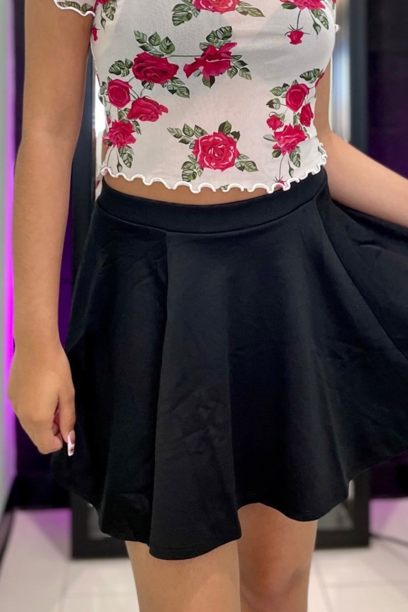 A person wearing a white cropped top with a red rose floral print and a black pleated mini skirt, shown from the chest down in an indoor setting with purple ambient lighting.