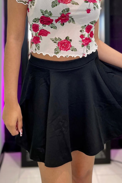 A person wearing a white cropped top with a red rose floral print and a black pleated mini skirt, shown from the chest down in an indoor setting with purple ambient lighting.