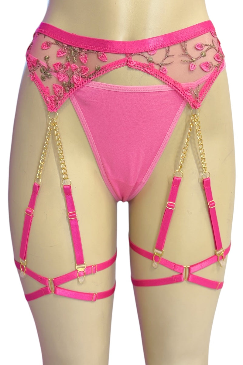 Bright pink lingerie set with a thong, sheer floral mesh side panels, and attached garter straps with gold-tone chains and buckles, displayed on a beige mannequin torso against a white background.