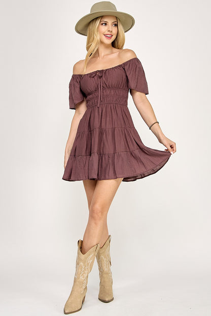 Woman wearing a brown off-shoulder dress with beige boots and a hat on a white background