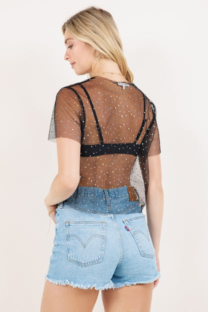 Woman wearing a sheer top with black bralette and black sequin polka dot overlay, paired with denim shorts on a white background.