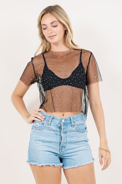Woman wearing a sheer top with black bralette and black sequin polka dot overlay, paired with denim shorts on a white background.
