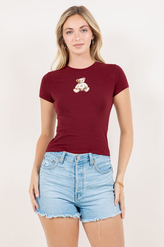 Woman wearing a burgundy t-shirt with a teddy bear design and denim shorts on a white background