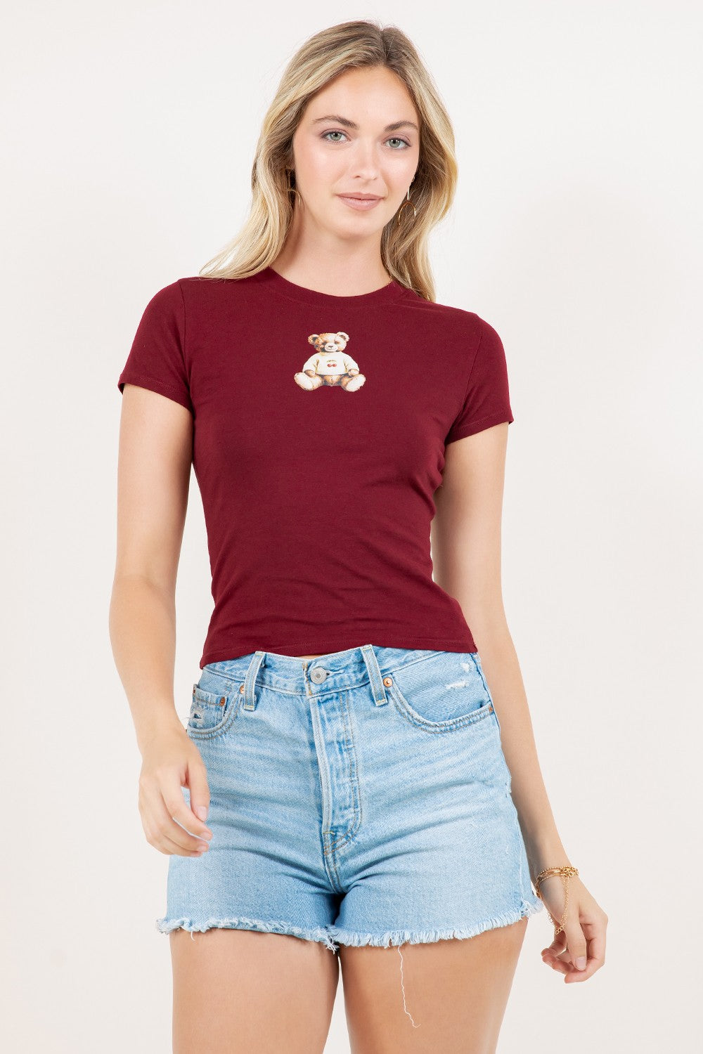 Woman wearing a burgundy t-shirt with a teddy bear graphic and denim shorts on a white background