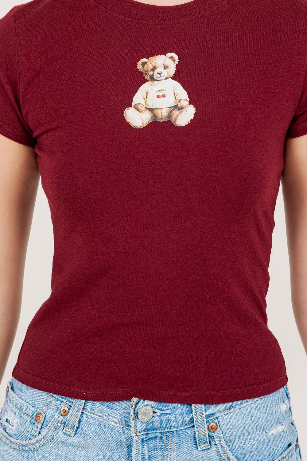 Burgundy t-shirt with a teddy bear design worn by a person, on a plain background