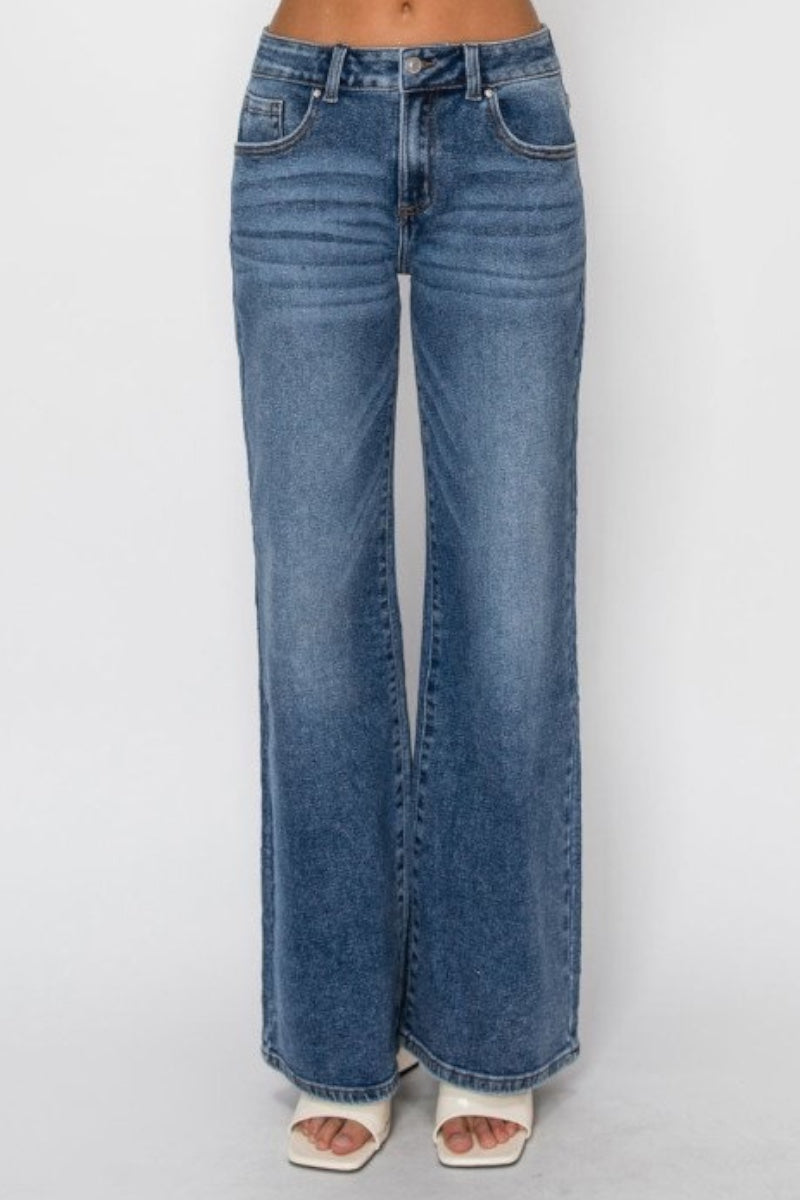 A person wearing dark-wash blue wide-leg denim jeans with white heeled sandals, shown from the waist down against a light gray studio background.