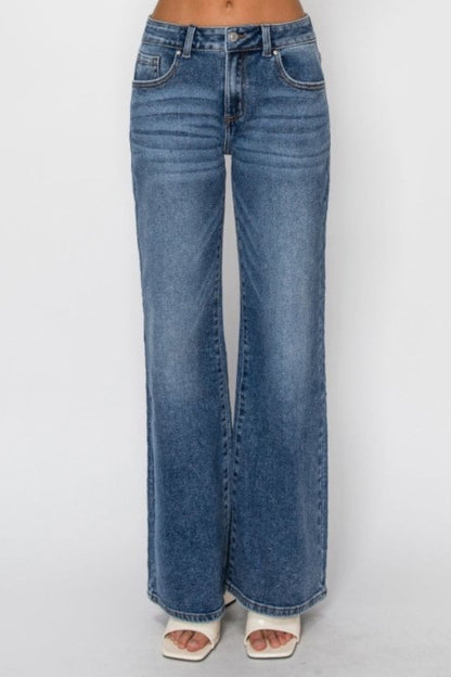 A person wearing dark-wash blue wide-leg denim jeans with white heeled sandals, shown from the waist down against a light gray studio background.