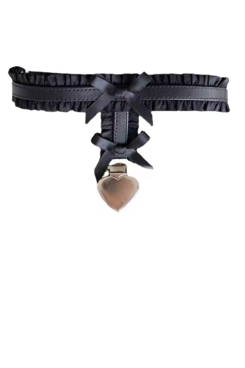 A black faux-leather leg garter featuring a ruffled edge, a central bow, lace-up detailing, and a dangling metallic heart-shaped charm, shown against a plain white background.