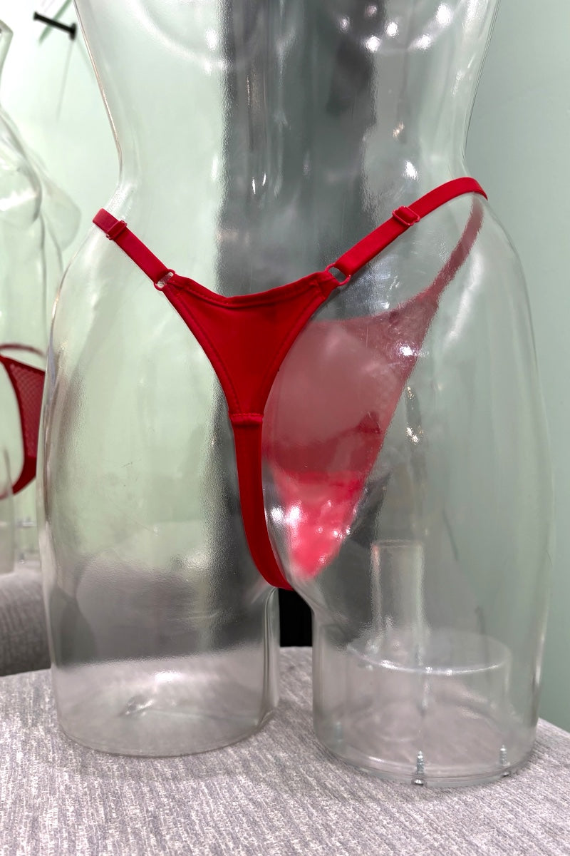 A red thong-style lingerie bottom displayed on a clear plastic torso mannequin, with a second mannequin partially visible at left and a pale green wall behind.