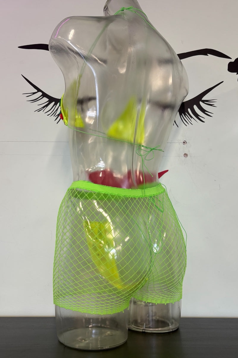 A clear plastic torso mannequin wearing neon green mesh shorts with a bright green waistband, displayed against a white wall with black eyelash graphics.