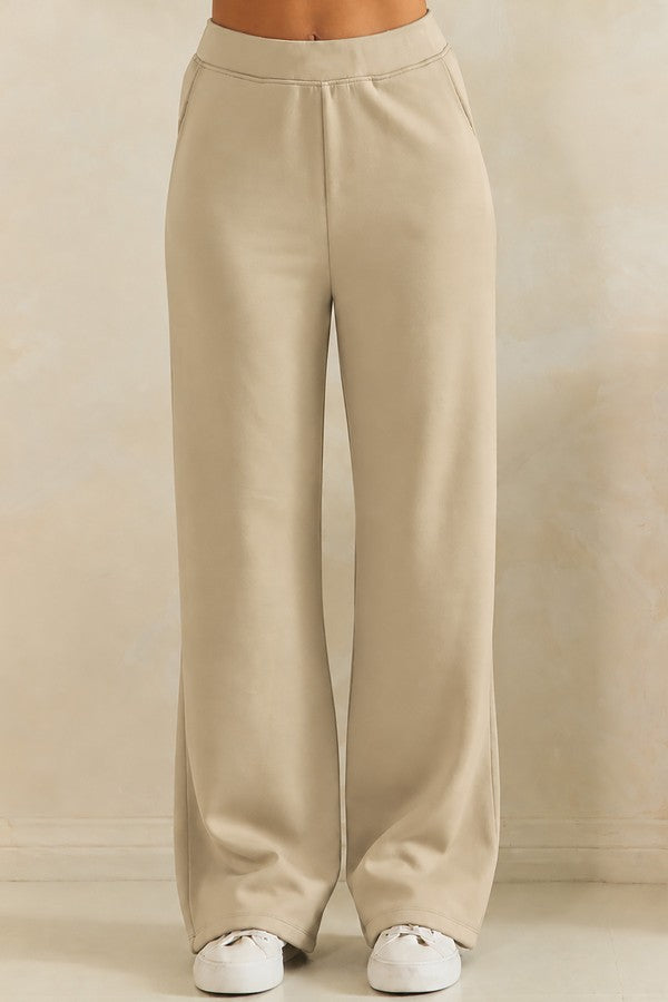 A person wearing high-waisted, wide-leg beige pants with white sneakers, shown from the waist down against a neutral beige studio background.