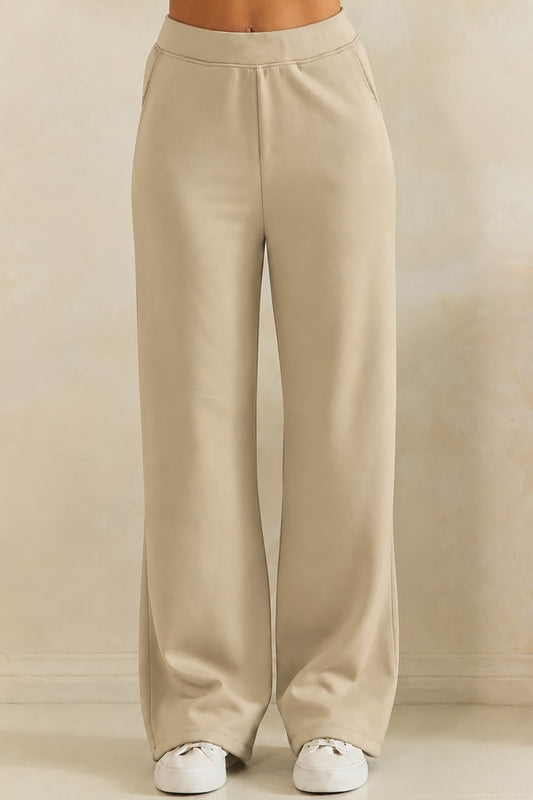 A person wearing high-waisted, wide-leg beige pants with white sneakers, shown from the waist down against a neutral beige studio background.
