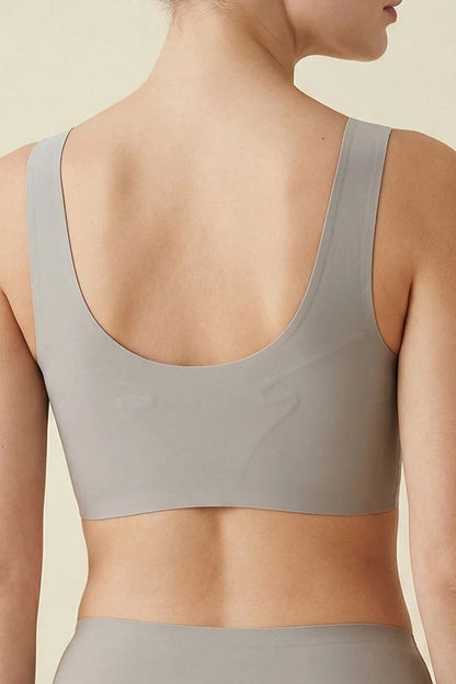 Rear view of a woman wearing a light taupe-gray bra with a wide scoop neckline and wide shoulder straps, paired with matching high-waisted bottoms against a warm beige background.