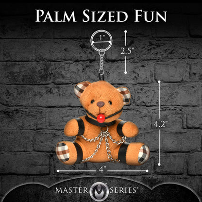 Palm-sized teddy bear keychain with measurement chart against a dark brick wall background.