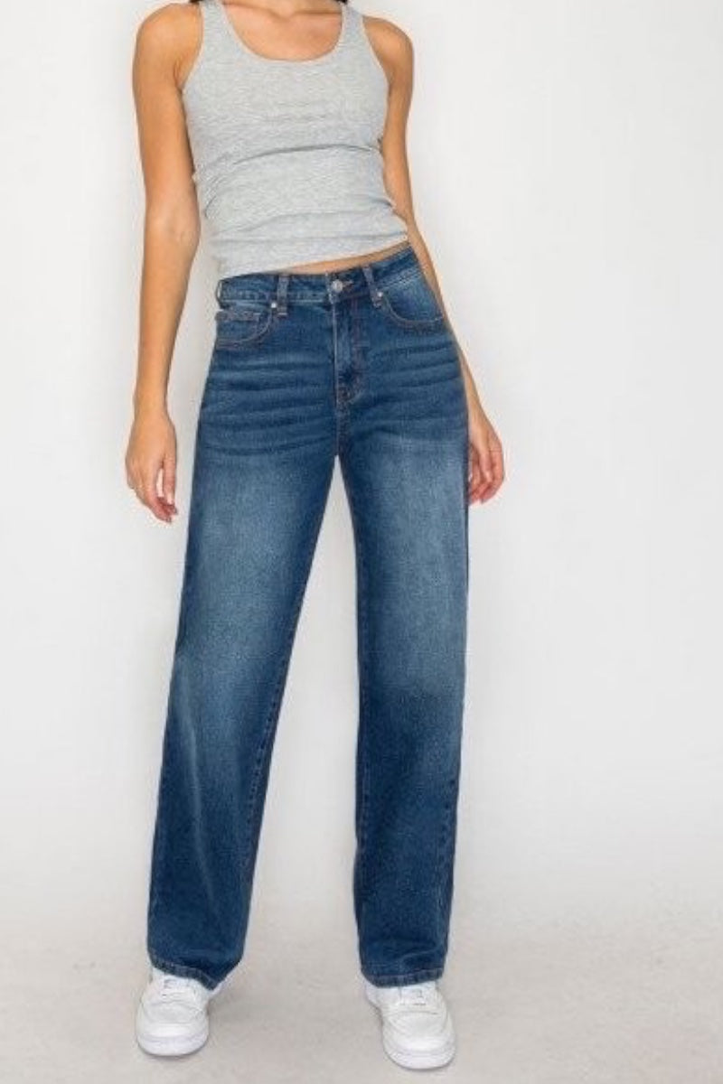 A model wearing a pair of high-waisted, wide-leg blue jeans with a gray ribbed tank top and white sneakers, standing against a plain white studio background.