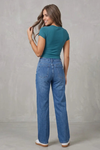 A woman is shown from the back wearing a teal short-sleeve top and high-waisted, wide-leg blue jeans against a textured gray studio backdrop.