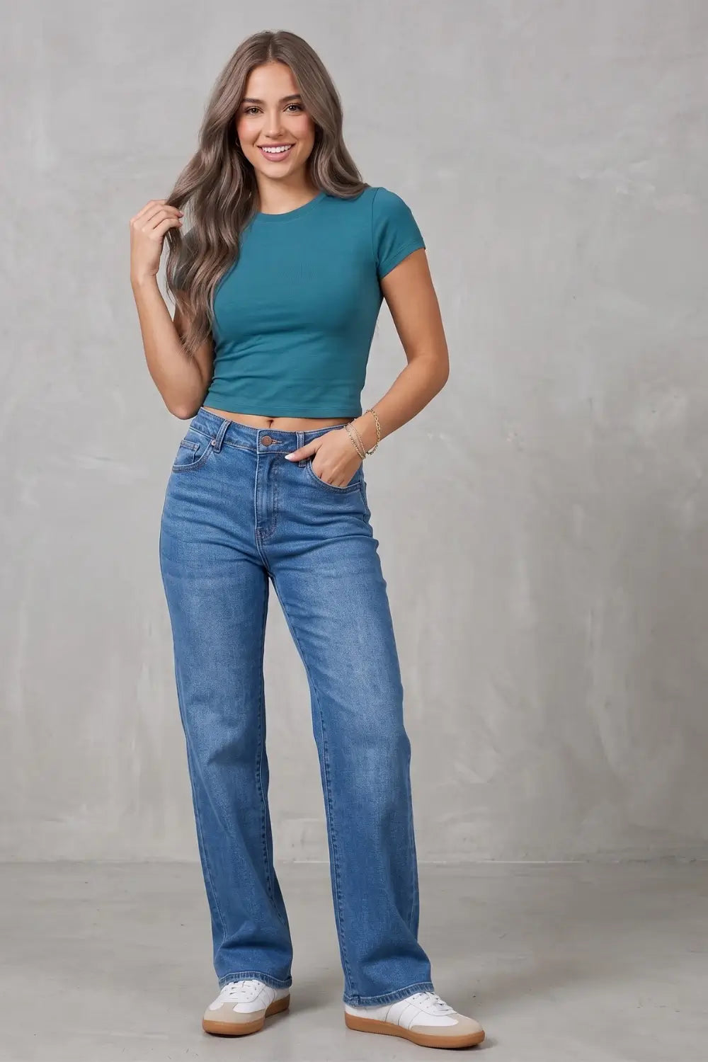 A woman modeling a teal short-sleeve crop top and high-waisted blue straight-leg jeans with white sneakers, standing against a textured gray studio backdrop.