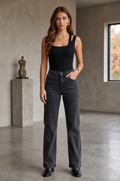 A woman modeling a black sleeveless top and dark gray wide-leg jeans in a minimalist studio setting with concrete walls and a window.