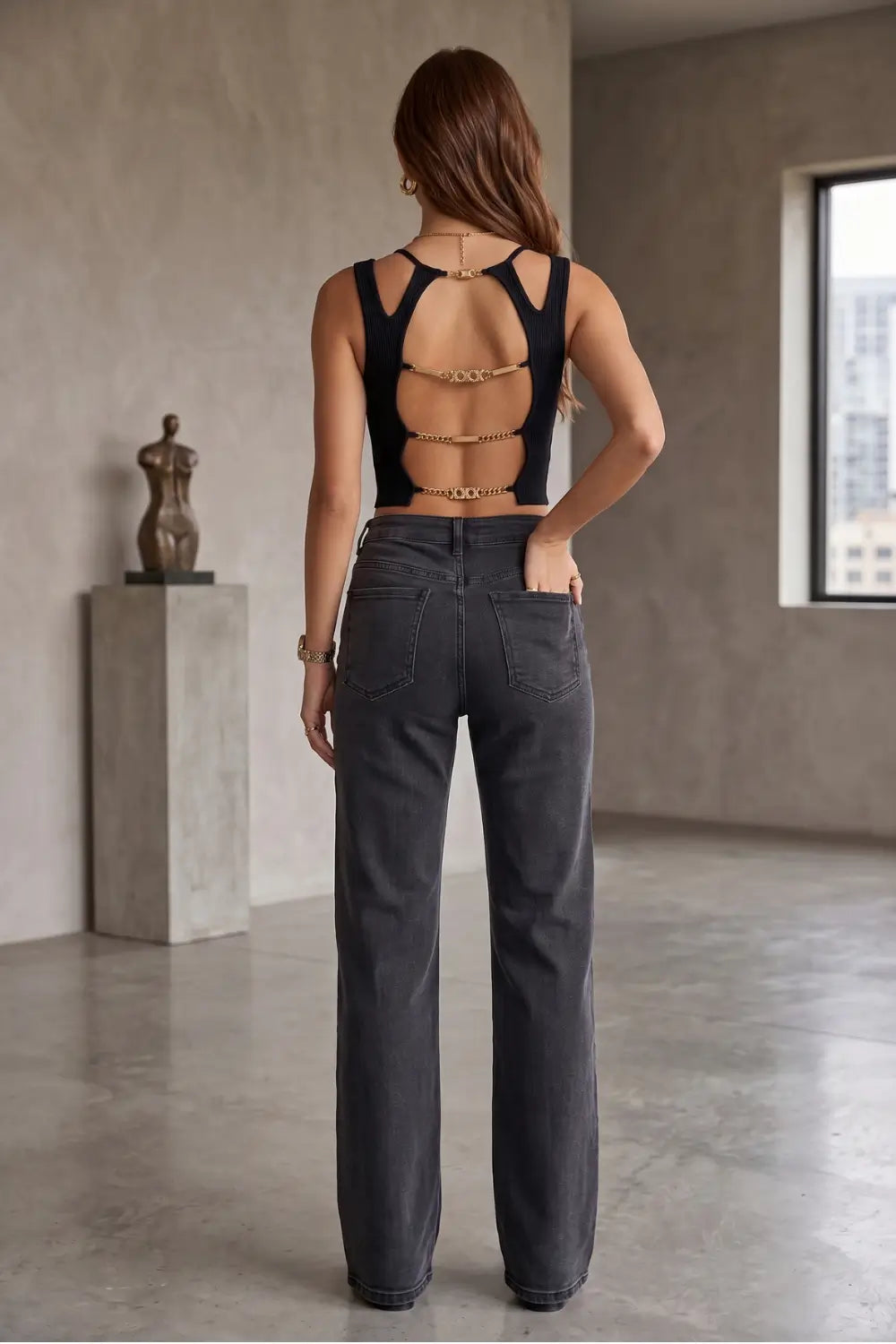 A woman stands with her back to the camera wearing a black strappy open-back top with gold chain accents and dark gray wide-leg jeans in a minimalist studio with concrete walls and a window.
