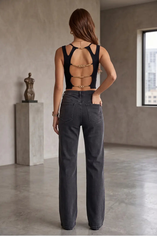 A woman stands with her back to the camera wearing a black strappy open-back top with gold chain accents and dark gray wide-leg jeans in a minimalist studio with concrete walls and a window.