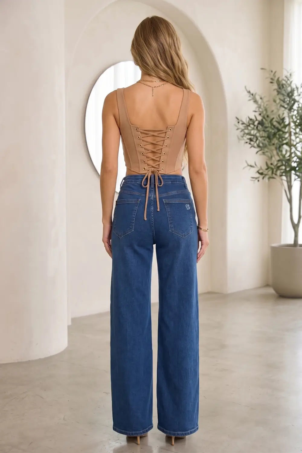 A woman viewed from behind wearing a beige lace-up back top and high-waisted blue wide-leg jeans in a bright, minimalist room with arches and a potted plant.