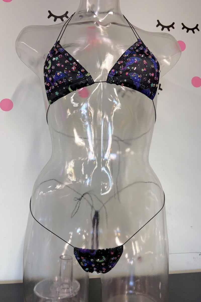 A two-piece bikini set with dark bats pattern, displayed on a clear plastic torso mannequin against a white wall with decorative eyelashes and pink dots.
