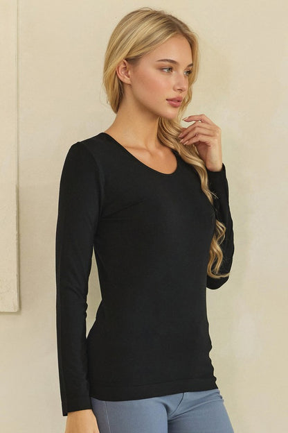 A blonde woman wearing a black long-sleeve scoop-neck knit top and light blue jeans, standing against a neutral beige wall.