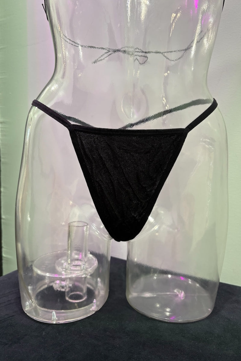 Clear plastic lingerie display form shaped like a torso, wearing black thong-style underwear, with a matching clear stand on a dark surface against a pale wall.
