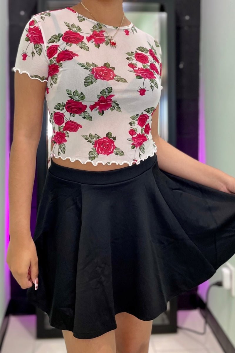 A woman wearing a white short-sleeve crop top with a red rose floral print and a black flared skirt, shown in a mirror selfie style.