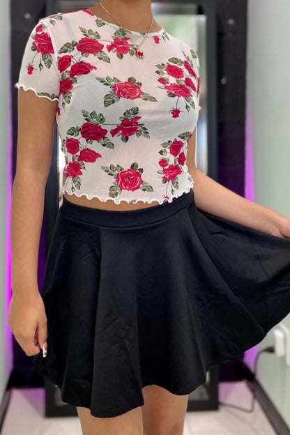 A woman wearing a white short-sleeve crop top with a red rose floral print and a black flared skirt, shown in a mirror selfie style.