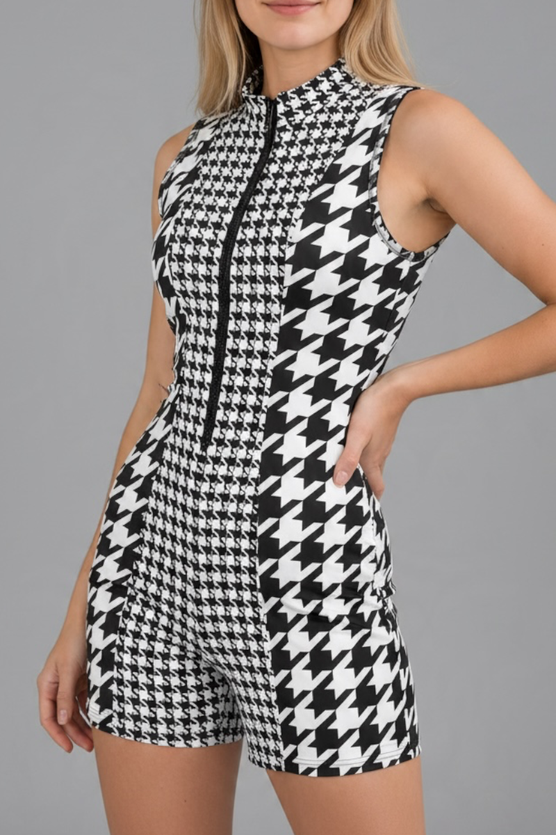 A sleeveless black and white houndstooth romper with a black front zipper, modeled against a solid gray background.