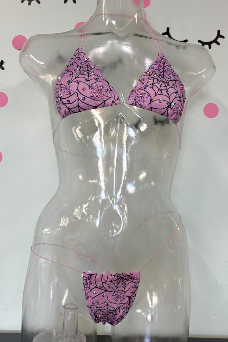 A pink and black spiderweb-patterned bikini set displayed on a transparent mannequin torso against a white wall with pink polka dots and black eyelash graphics.