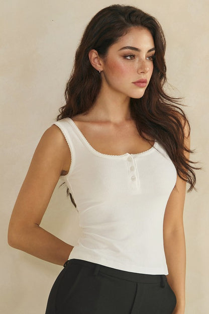 A woman modeling a fitted white sleeveless henley top with a scoop neckline and small front buttons, paired with black pants, against a plain beige studio background.